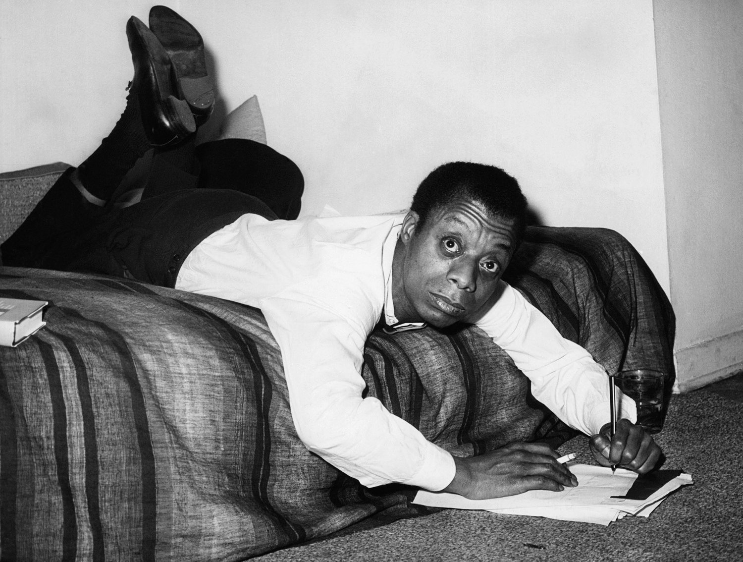 Black and white photo of a man lying down looking up from the edge of a bed while writing