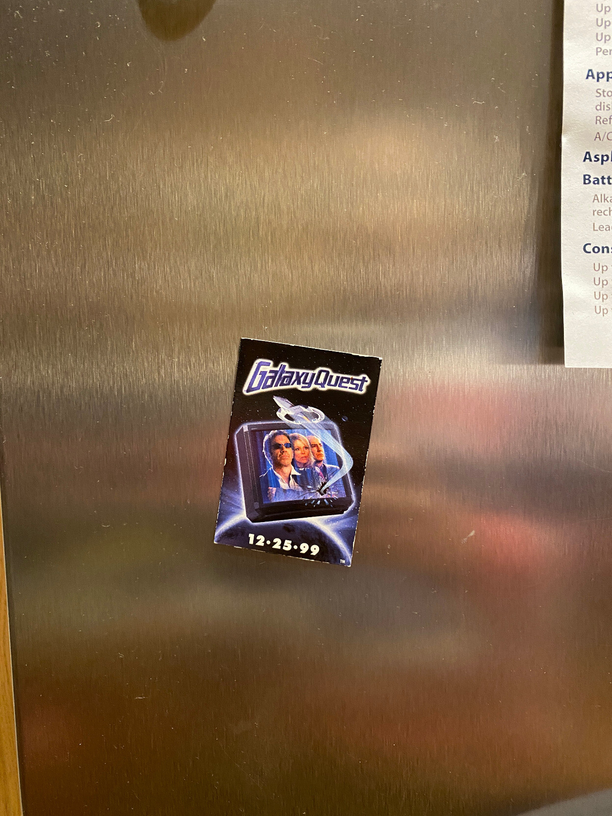 A stainless steel refrigerator door with a Galaxy Quest magnet stuck to it. The magnet is rectangular, two inches by three inches, portrait orientation. It's black with the Galaxy Quest logo at the top and an image of the three lead actors on a TV screen that is floating in outer space below the logo. An image of the spaceship they flew is zooming out of a crack in the TV screen and flying up between the TV and the logo at the top. At the bottom it says the release date, 12-25-99.