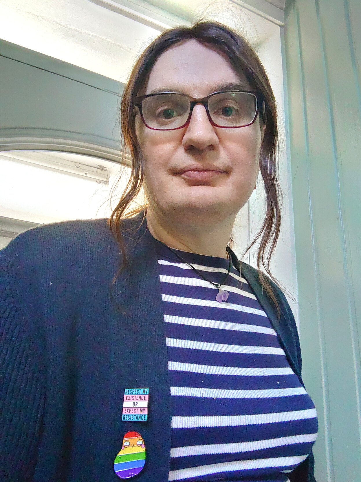 The same person presenting femininely, beardless, with long brunette hair pulled partially into a pony tail, leaving a couple of locks free to frame the face. They're wearing a navy and white striped top, a fluorite pendant, a navy cardigan, and two pins-- a trans flag colour one saying "respect my existence or expect my resistance", and a pride flag colour one in the shape of a cartoon potato. You can see that transition has given the person softer features and a noticeable bust.