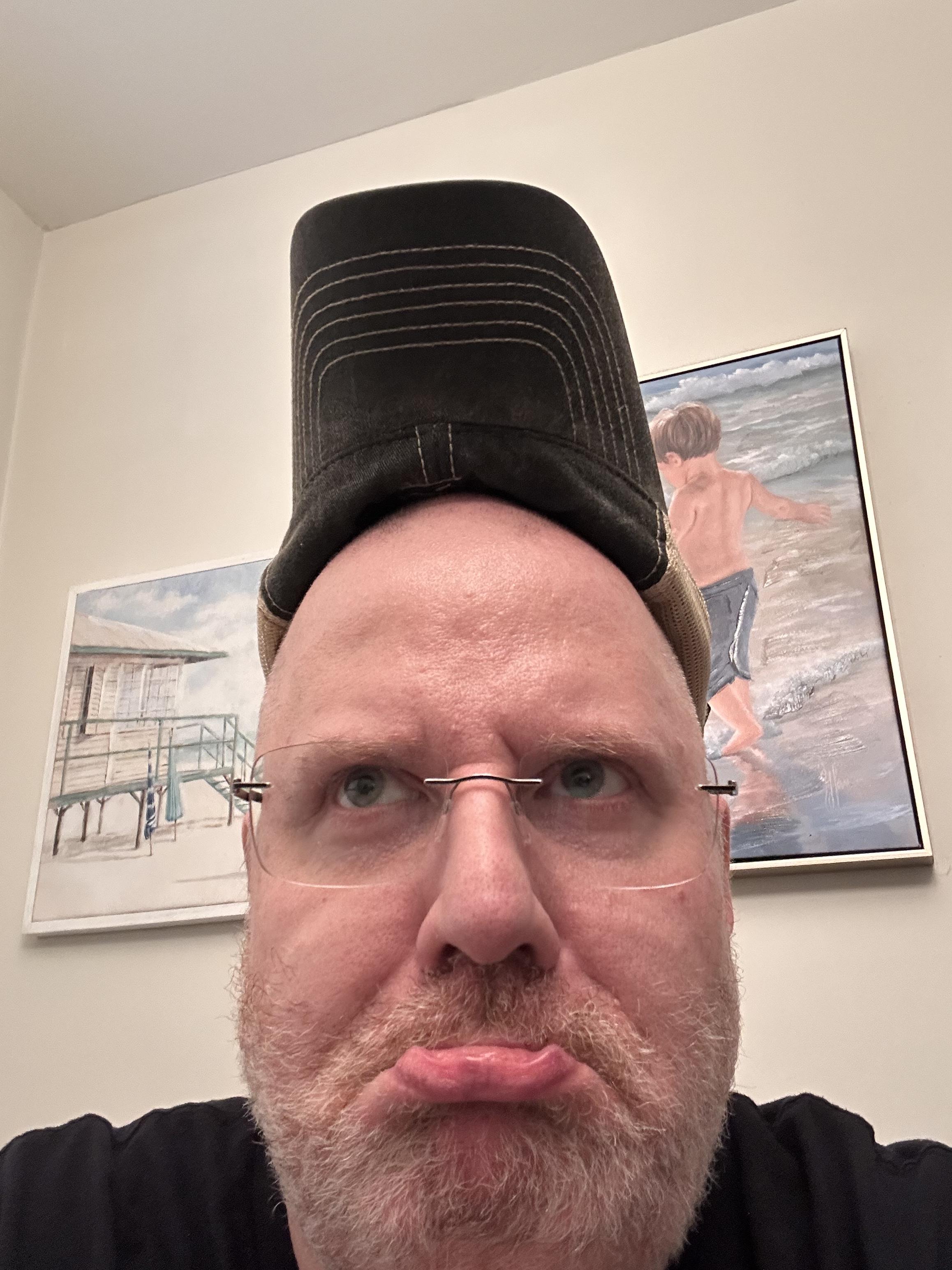 A selfie of a middle aged man, frowning dramatically while wearing his baseball hat “rally style”. 