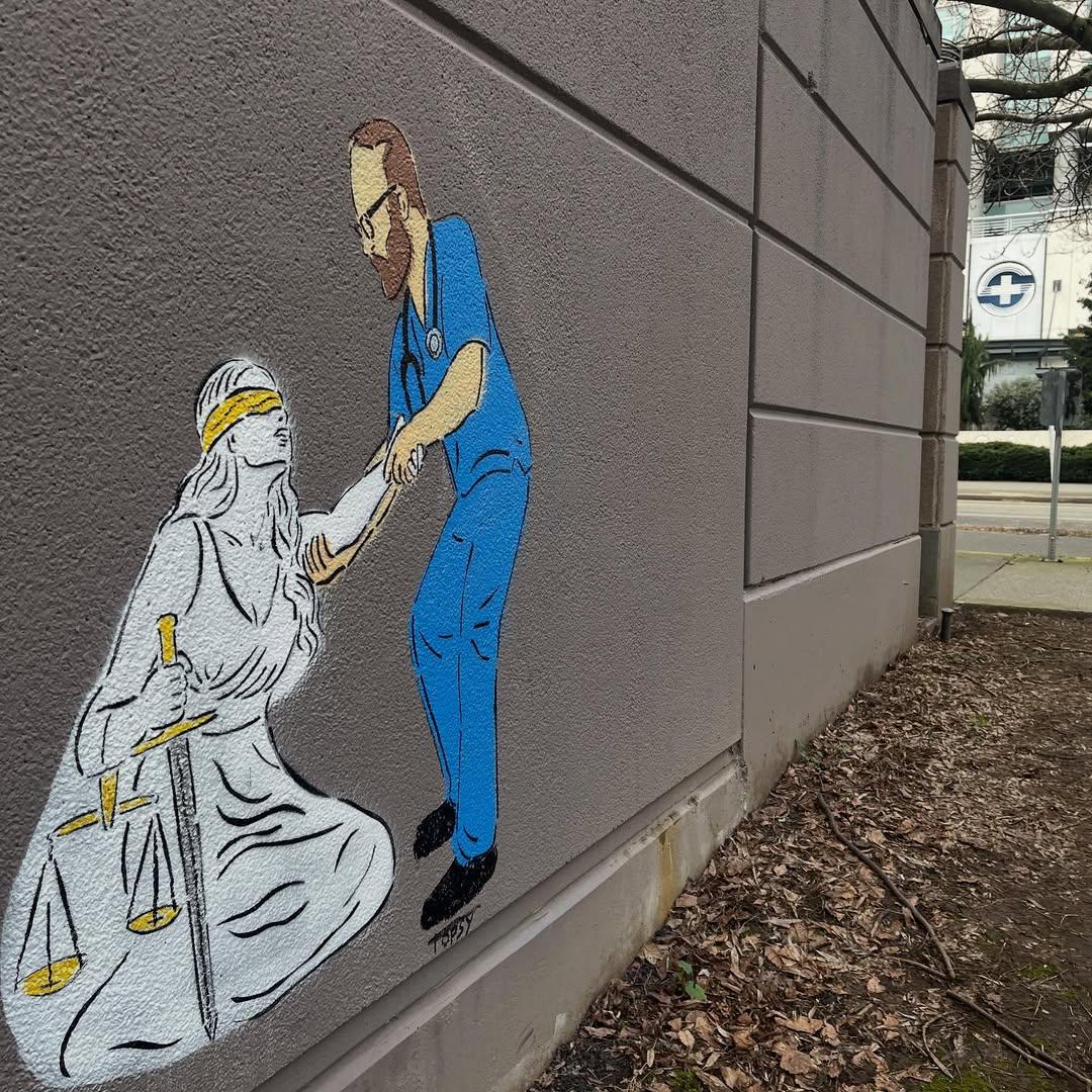 Street art by Topsy (topsy_paints) in Seattle, USA, painted on the corner of a gray building wall: a nurse/doctor in bright blue scrubs with a stethoscope leans down to lift a kneeling Lady Justice in a white robe, her eyes covered with a gold blindfold, holding a sword while the scales of justice hang at her side; the wider photo shows the mural in its urban setting with sidewalk, trees, and street in the background.