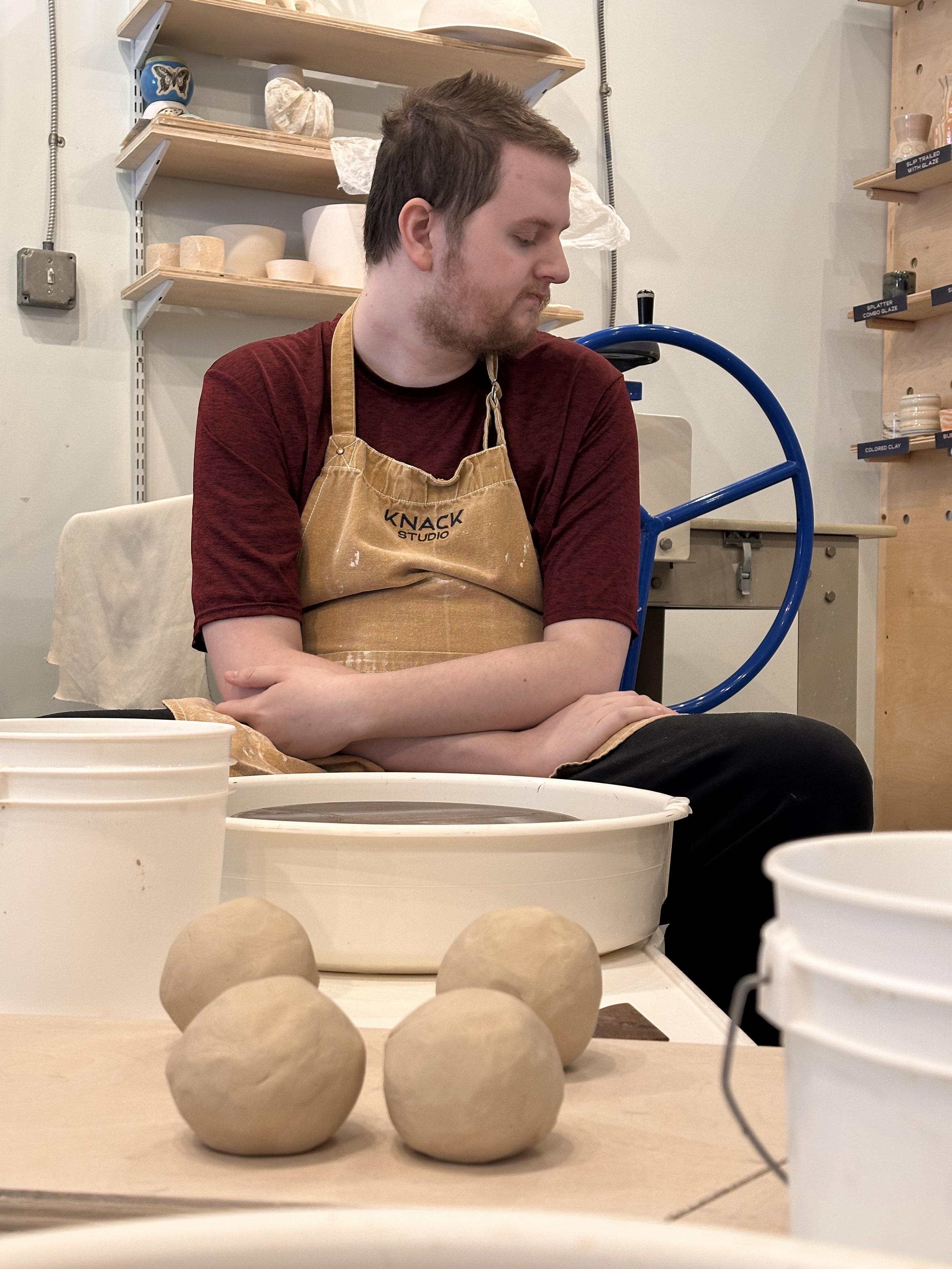 My son in an apron, sitting at a pottery wheel, watching a demonstration, which is happening out of frame. He’s a tall young man with short brown hair and a scruffy beard. 