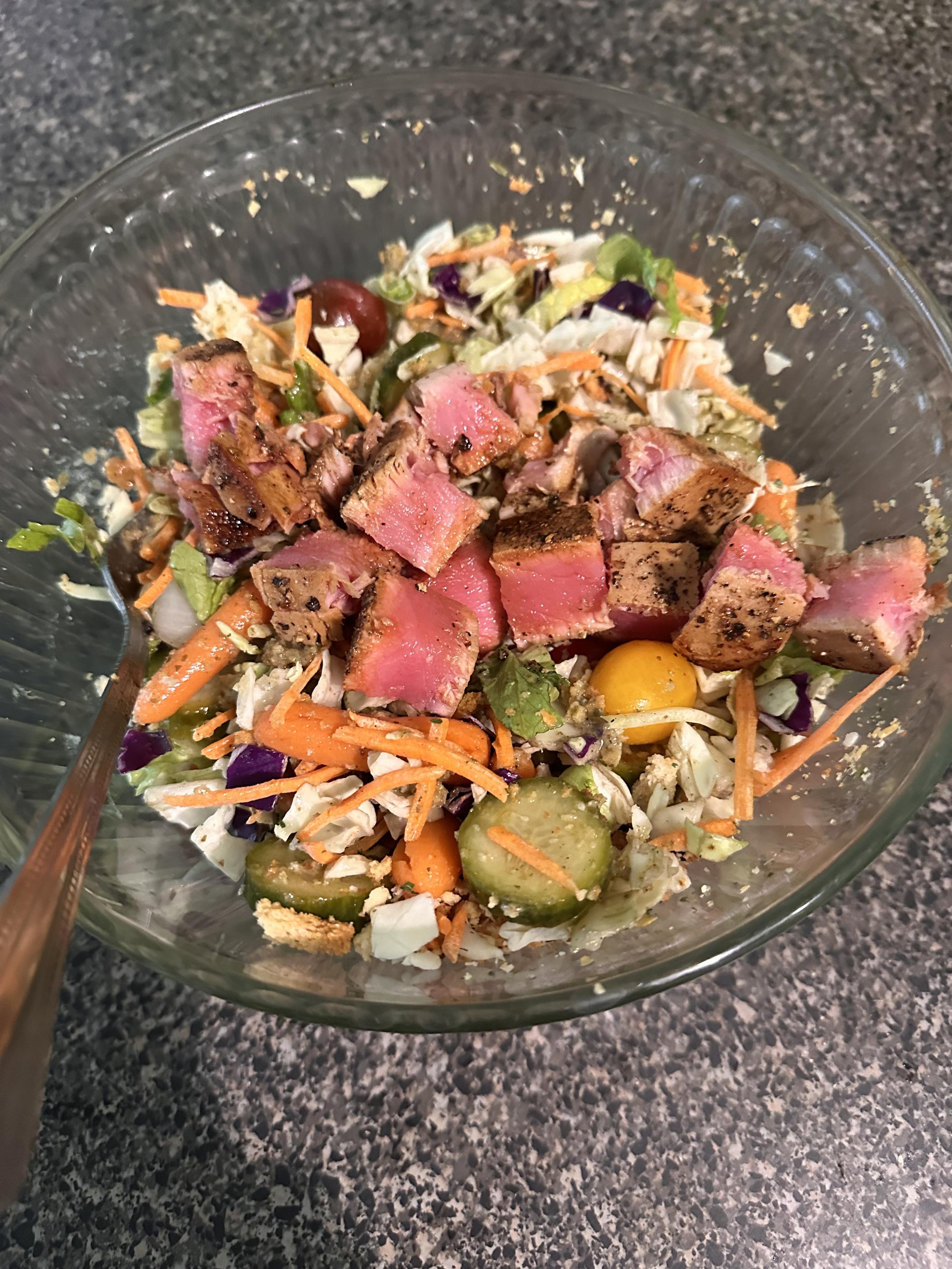 A salad topped with rare seared tuna cut into cubes, pickled cucumbers, carrots and onions in a glass bowl. 
